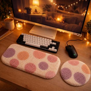 Pode incluir: Um apoio de pulso retangular para teclado e um mouse pad circular, ambos com uma textura tufada na cor creme e um padrão de grandes bolinhas em tons suaves de lavanda, pêssego e rosa claro, dispostos sobre uma superfície de madeira.
