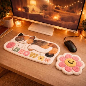 May include: A cat-shaped rug with the word "MEOW" and a flower-shaped coaster with a smiling face. The rug features a calico cat design in brown, orange, and black. The coaster is pink and yellow. The items are on a wooden desk.