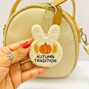 May include: A cream-colored, punch needle bunny-shaped keychain featuring an embroidered orange pumpkin flanked by two yellow wheat stalks. The text "AUTUMN TRADITION" is stitched in black thread below the pumpkin. The keychain hangs from a silver ring.