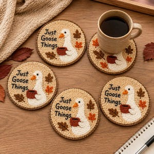 Könnte beinhalten: Ein Set aus vier runden, beigen Häkeluntersetzern mit einer weißen Gans, die eine braune Tasse hält, umgeben von kleinen braunen und orangefarbenen Herbstblättern. Der Text "Just Goose Things." ist auf jedem Untersetzer in Schwarz gestickt.