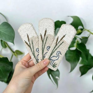 以下が含まれることがあります： クリーム色の背景に、背の高い緑の葦と青いサギが刺繍された、3枚の長方形のパンチニードルパッチ。質感のある布地は上端が丸みを帯びており、一貫した詳細な刺繍パターンが施されています。