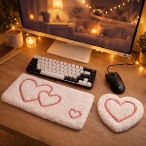 May include: A cozy desk setup featuring a white and black keyboard, a black mouse, and a white heart-shaped mousepad and a rectangular heart-themed mat. The mat and mousepad have pink heart embroidery. The desk is made of wood.