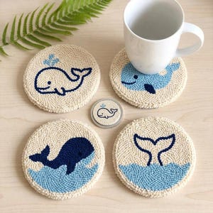 Puede incluir: Juego de cuatro posavasos redondos con diseños de ballenas azules. Los posavasos son de color blanco roto. Uno tiene una ballena que arroja agua, otro una ballena nadando y un tercero muestra la cola de una ballena. Una taza blanca está sobre uno de ellos.