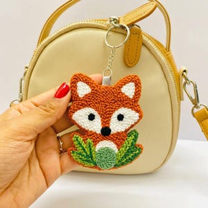 May include: A fox-shaped keychain featuring orange, white, and black yarn embroidery with green leaf accents at the base. The fox has pointed ears, a white face patch, and black eyes and nose, attached to a silver metal ring and chain.