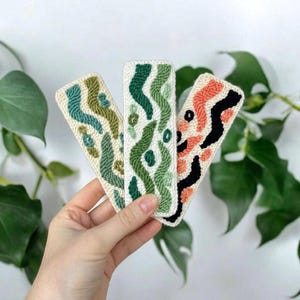 May include: Three rectangular punch needle bookmarks featuring textured yarn designs. Two bookmarks display wavy green and olive patterns on a cream background, while the third features coral and black abstract shapes on a cream background.