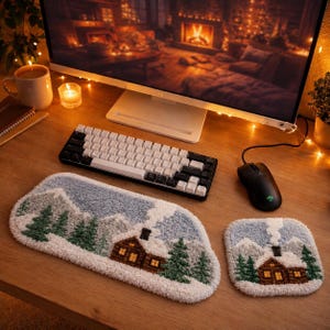 Könnte beinhalten: Eine rechteckige Tastatur-Handballenauflage und ein quadratisches Mauspad mit getuftetem Winterlandschaftsmotiv, das eine verschneite Blockhütte, grüne Tannen und Berge vor einem hellblauen Himmel auf einem Holztisch zeigt.