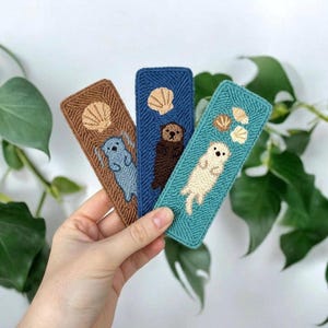 May include: Three rectangular embroidered bookmarks featuring sea otters and seashells. The left bookmark is brown with a blue otter, the center is blue with a dark brown otter, and the right is teal with a cream-colored otter and three shells.