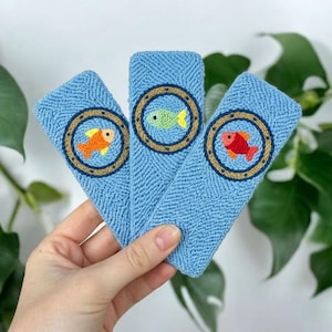 May include: Three rectangular light blue punch needle hair clips, each featuring a circular brown porthole design. Inside the portholes are embroidered fish in orange, light green, and red, with small black eyes and yellow fins.