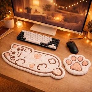 May include: A white and brown cat-shaped desk mat and a paw-shaped mouse pad sit on a wooden desk. A black and white keyboard and a black mouse are also on the desk. The desk is in front of a computer monitor.