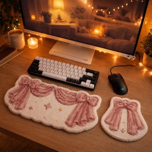 Può includere: Un tappetino rettangolare per tastiera e un tappetino per mouse quadrato più piccolo, entrambi con una superficie trapuntata color crema con disegni di fiocchi rosa e piccoli accenti a stella ricamati. I tappetini sono posizionati su una scrivania in legno.