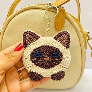 Può includere: Un portachiavi a forma di gatto realizzato in filato color crema e marrone scuro, con occhi in plastica blu e baffi in filo marrone. Il portachiavi presenta un anello e una catenella in metallo, con una texture lavorata a mano.