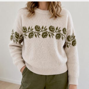 May include: A cream-coloured knitted jumper with a crew neck and long sleeves. The jumper has an olive green floral pattern across the chest and on the upper sleeves. The material is soft and textured.