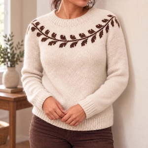 Könnte beinhalten: Ein cremefarbener Langarm-Strickpullover mit Rundhalsausschnitt und einem dunkelbraunen Blattmuster an der Passe. Der Pullover verfügt über gerippte Bündchen an Ärmeln und Saum und ist aus einem weichen, strukturierten Garnmaterial gefertigt.