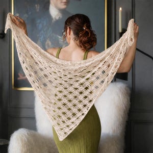 May include: A cream-colored, open-weave shawl with a geometric pattern. The triangular shawl is held up, showcasing its delicate design. The shawl is made of a soft, lightweight material, perfect for layering. The shawl is a versatile accessory.