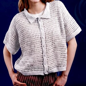 May include: A light gray, short-sleeved crochet shirt with a collared neckline and button-down front. The shirt has a loose, open weave design. The person is wearing a white t-shirt underneath and striped pants.
