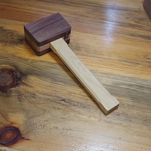 May include: A wooden mallet featuring a dark brown rectangular head with a lighter brown center stripe and a long, light-colored handle. The head displays a small, rectangular maker's mark stamped on the side near the handle connection point.