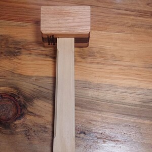 May include: A wooden mallet featuring a rectangular head constructed from light-toned wood with a dark wood accent layer. The handle is made of a smooth, light-colored wood with a rectangular profile and beveled edges, extending from the mallet head.