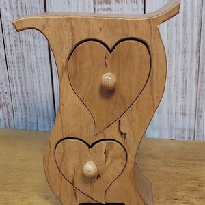 May include: Wooden jewelry box with two heart-shaped drawers and small wooden knobs. The box has a curved, owl-like shape and is made of light-colored wood. The drawers are carved with heart shapes.