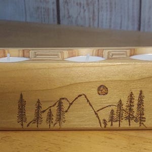 May include: Wooden candle holder with three tea lights. The light-colored wood has a mountain and tree design etched into the front. The candle holder is rectangular with a slightly angled front.