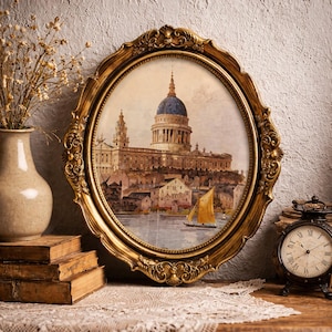 May include: An oval, gold-colored ornate picture frame with a watercolor painting of a cathedral with a blue dome. A small sailboat is on the water. A vase with dried flowers, old books, and a clock are nearby.