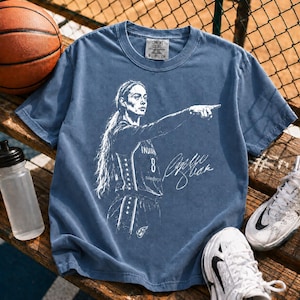 May include: A blue short-sleeve t-shirt featuring a white graphic illustration of a basketball player pointing, with the text "INDIANA 8" and a signature. The shirt has a Comfort Colors label at the inner neckline and a crew neck design.