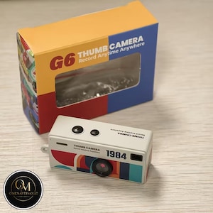 Puede incluir: Una cámara en miniatura blanca con un diseño retro, con el texto "THUMB CAMERA" y "1984". La cámara tiene un diseño de rayas coloridas y dos elementos negros con forma de lente. La caja es naranja, roja y azul con el texto "G6 THUMB CAMERA Record Anytime Anywhere".