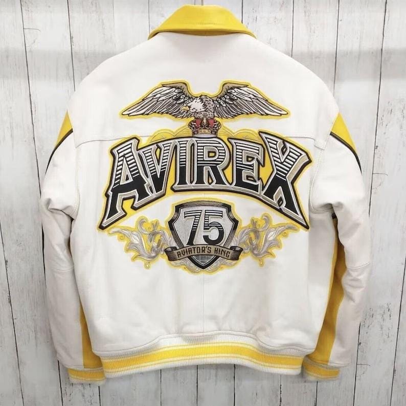 May include: White leather jacket with yellow detailing, featuring the text "AVIREX 75 AVIATOR'S KING". Includes an eagle emblem and a yellow collar. A classic bomber jacket design.