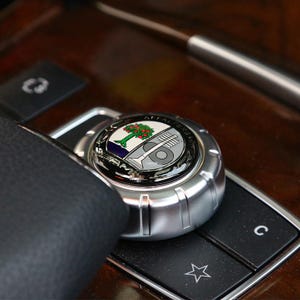 May include: A silver-colored car control knob with a circular emblem featuring a tree, musical instrument, and text. The knob is set on a dark wooden surface with other black buttons.
