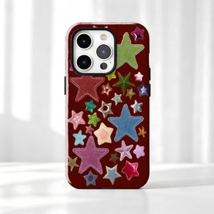 May include: A dark red smartphone case featuring a scattered pattern of various star-shaped icons. The stars appear in diverse textures, finishes, and colors, including denim blue, glittery green, metallic gold, pink, and light blue, creating a collage.