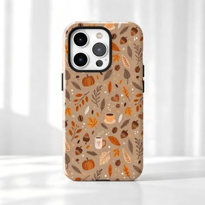 May include: A smartphone case with a tan background featuring a repeating pattern of orange pumpkins, brown acorns, various autumn leaves in shades of orange and brown, and small mugs with steam, accented by tiny white dots throughout the design.