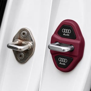 May include: A comparison of a rusty car door latch and a new burgundy Audi door latch cover. The new cover has the Audi logo and the word "Audi" in white on a black background.