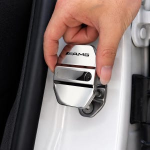 Op de afbeelding: Een verchroomde auto deurslotafdekking met het AMG-logo in zwart. De afdekking wordt op een witte autodeur gemonteerd. De rechthoekige afdekking heeft een uitsparing en is gemaakt van glanzend metaal.