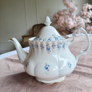 Puede incluir: Una tetera de porcelana blanca con cuerpo festoneado y tapa a juego con un remate puntiagudo. La tetera está decorada con un delicado patrón de pequeñas flores nomeolvides azules y moradas, y acentuada con un fino borde dorado en los bordes.