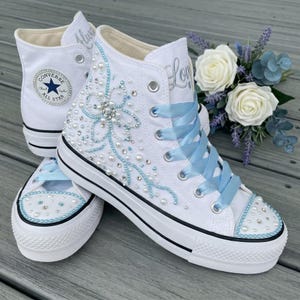 Peut inclure: Baskets montantes blanches avec des lacets en ruban bleu clair et des ornements. Les baskets présentent des accents de perles et de strass, avec le mot "Love" en script argenté. Le logo Converse est visible sur le côté.