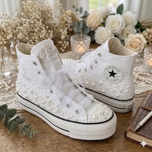 Könnte beinhalten: Weiße High-Top-Sneaker, verziert mit Perlen und Schleifenbändern. Die Sneaker haben eine schwarze Sohle und einen Converse All Star-Aufnäher. Der linke Schuh hat die Initialen "J D" und das Datum "05.18.24" auf der Seite gestickt.