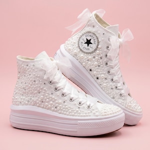May include: A pair of white high-top platform sneakers featuring satin ribbon laces, sheer organza bows, and an exterior embellished with scattered white pearls and clear rhinestones. The side displays a circular Converse All Star logo patch.