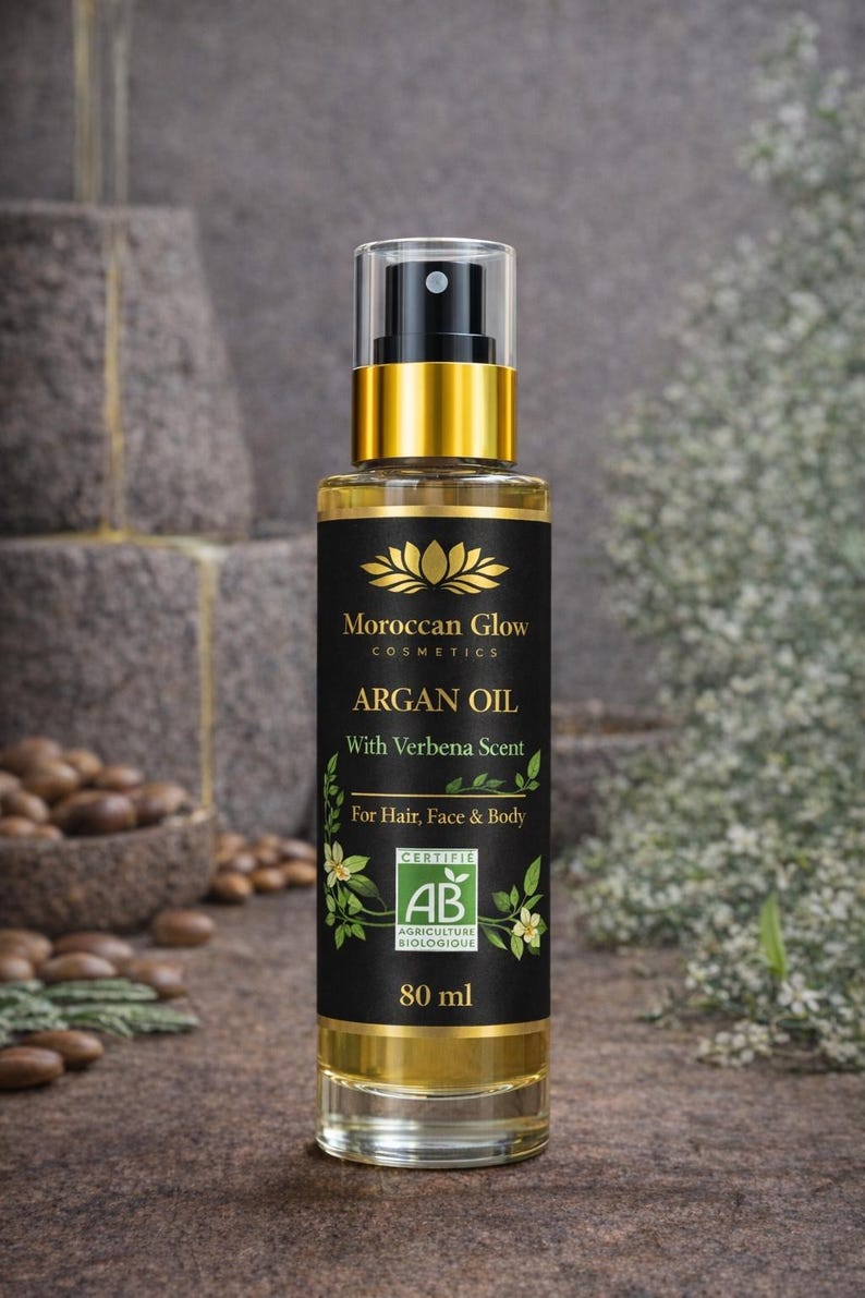 Puede incluir: Una botella de vidrio transparente de Aceite de Arg&aacute;n Moroccan Glow con una etiqueta negra y dorada. La etiqueta dice "ARGAN OIL With Verbena Scent For Hair, Face & Body" e incluye un logotipo de certificaci&oacute;n. La botella contiene 80 ml de aceite.