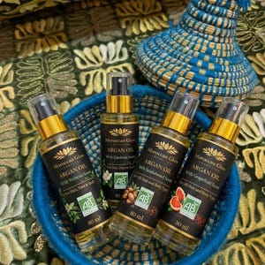 Puede incluir: Una cesta azul tejida contiene cuatro botellas de 80 ml de aceite de arg&aacute;n Moroccan Glow con diferentes aromas. Las botellas tienen etiquetas negras con detalles dorados. Un sombrero tejido azul y amarillo est&aacute; en el fondo.