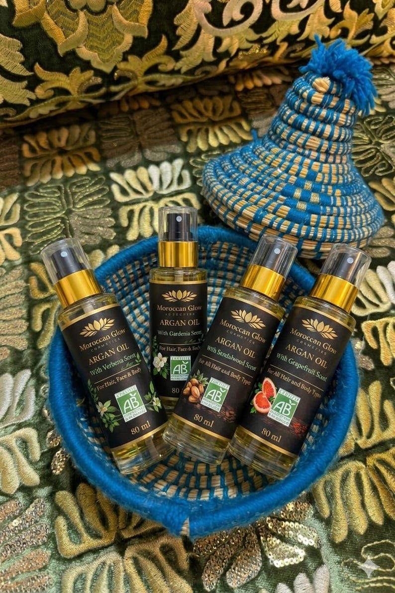 Puede incluir: Una cesta azul tejida contiene cuatro botellas de 80 ml de aceite de arg&aacute;n Moroccan Glow con diferentes aromas. Las botellas tienen etiquetas negras con detalles dorados. Un sombrero tejido azul y amarillo est&aacute; en el fondo.