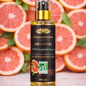 Puede incluir: Una botella de aceite de arg&aacute;n Moroccan Glow con aroma a pomelo, para cabello y cuerpo, se muestra sobre un fondo de rodajas de pomelo. La botella es de 80 ml y tiene una etiqueta dorada y negra con la certificaci&oacute;n AB Agriculture Biologique.