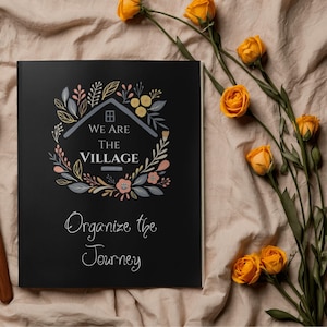 May include: A black journal with the text "We Are The Village" and "Organize the Journey" in white script. The cover features a floral wreath design with a house graphic. Yellow roses are arranged on a beige fabric background.