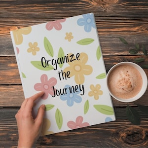 Peut inclure: Un cahier blanc avec le texte "Organize the Journey" et des illustrations florales aux couleurs pastel. Une tasse de café est posée à côté du cahier sur une surface en bois.