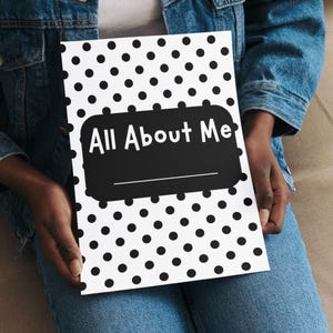 May include: A black and white notebook with a polka dot pattern. The notebook has the text "All About Me" in white on a black rectangular label. The notebook is held by a person wearing a denim jacket.