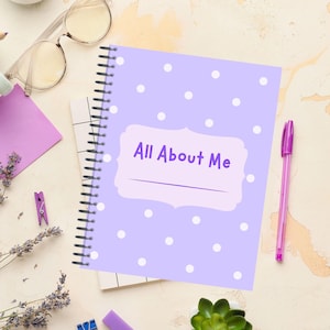 May include: A spiral-bound notebook with a lavender cover featuring white polka dots. The cover has a light purple label with the text "All About Me". A purple pen rests beside the notebook, along with eyeglasses and other stationery.
