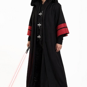Puede incluir: Larga túnica negra con capucha y detalles rojos en las mangas. La túnica tiene un diseño largo con botones en la parte delantera. Se sostiene un sable de luz rojo. Es un disfraz de cosplay.