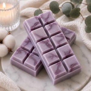 May include: Two rectangular wax melt bars in a soft lavender shade with a marbled purple swirl pattern. Each bar is divided into ten individual square segments. The wax melts are positioned on a round white marble surface with a smooth finish.