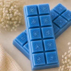 May include: Two rectangular blue wax melt bars, each divided into ten square segments. The surface of each segment features a textured, crystalline pattern that resembles small, clustered droplets or beads in a lighter shade of blue.