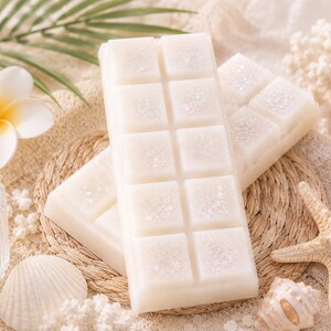 May include: Two rectangular white wax melt bars featuring a segmented grid pattern and a light dusting of iridescent glitter. The wax bars are positioned on a round woven mat, accompanied by a white flower, a starfish, and a small seashell.