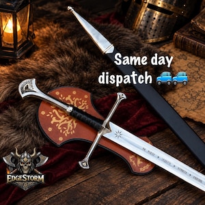 May include: A silver-toned metal sword with a black leather-wrapped grip and a crossguard, featuring an engraved star and runic inscriptions on the blade. It is accompanied by a black scabbard with silver accents and a brown leather display plaque.