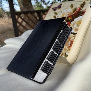 May include: A black leather-bound journal with bamboo leaf embossing, open to reveal labeled tabs. A second book with floral and butterfly illustrations is visible in the background. The journal's tabs are labeled with text.